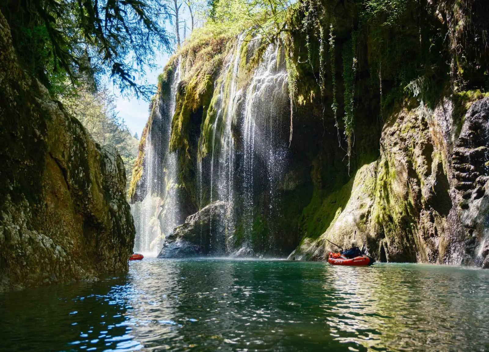 Kayakers on turquoise water beneath mossy waterfalls in the Gorges du Fier canyon